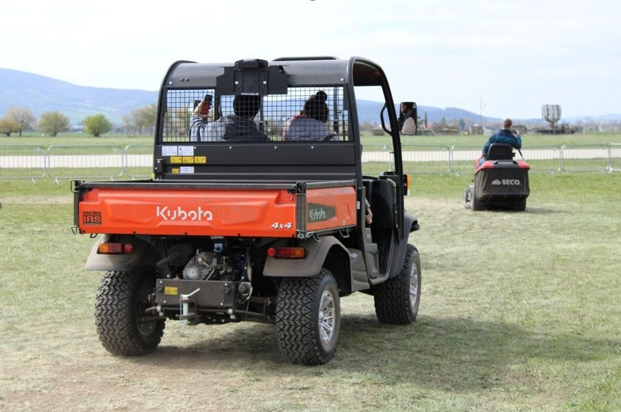 Úžitkové vozidlo Kubota s pasažiermi jazdiacimi po trávnatej ploche na záhradníckej akcii, s ďalšími poľnohospodárskymi strojmi v pozadí.